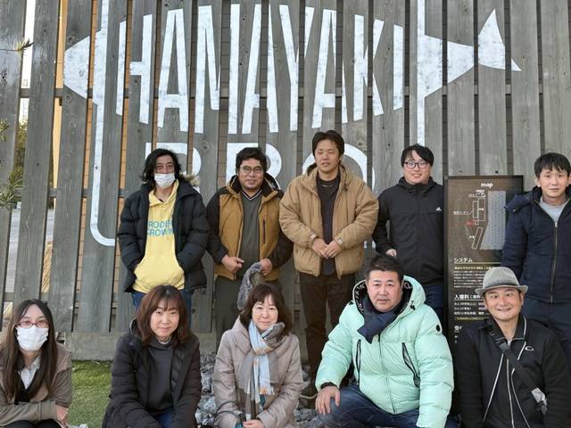親睦会のメンバーが外で集合写真を撮影している様子。背景に木製のフェンスと大きな文字が見える。