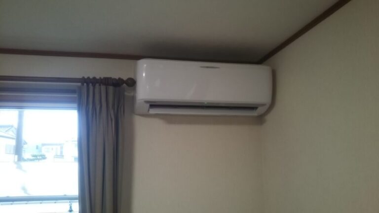 壁掛け型の白いエアコンが部屋の壁に取り付けられ、窓際にカーテンがある様子